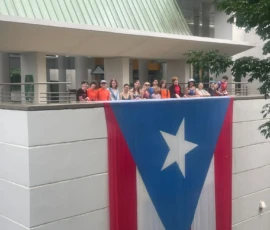 A group of people stands on a balcony above a large Puerto Rican flag hanging from the white building's exterior.