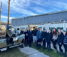 A group of twelve people stands beside a metal boat and a white fence outdoors on a sunny day, wearing dark clothing and boots.