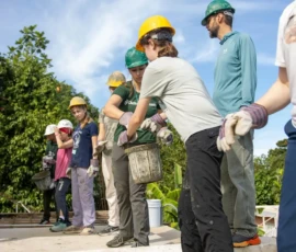 A group of people wearing hard hats and gloves pass a bucket down a line outdoors on a sunny day, participating in a construction or teamwork activity.