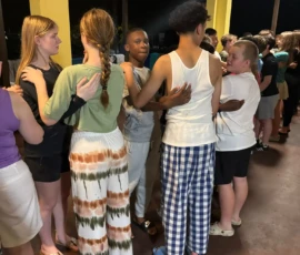 A group of young people stand in two lines facing each other, placing their hands on each other's shoulders, on a covered porch at night.
