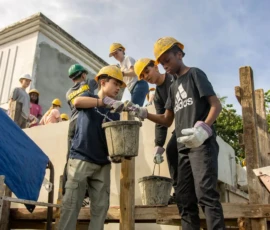 A group of people wearing yellow hard hats passes buckets of cement while working together on a construction site outdoors.