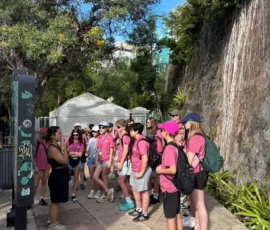 A group of people in matching pink shirts stand outdoors, listening to a tour guide near a stone wall and greenery on a sunny day.