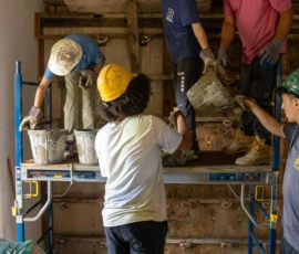 Five people in construction gear work together on scaffolding, passing buckets of material inside a room with tiled floors and unfinished walls and ceiling.
