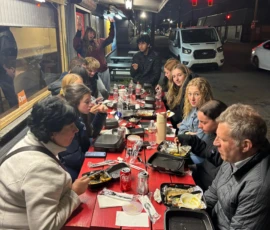 A group of people sit around a red picnic table outdoors at night, eating takeout food and drinking soda under overhead lights.
