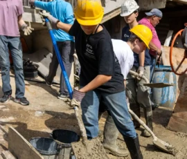 A group of people in hard hats and work gloves pour and spread wet concrete into a wooden frame at a construction site.
