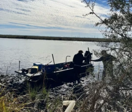 Two people stand next to a small motorboat stuck in muddy terrain along the bank of a river under a partly cloudy sky.