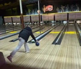 A person in a dark jacket and light pants is bowling, mid-throw, in a bowling alley with several lanes and pins visible in the distance.