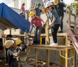 A group of people wearing hard hats and gloves work together on scaffolding to build or repair a structure under a partly cloudy sky.