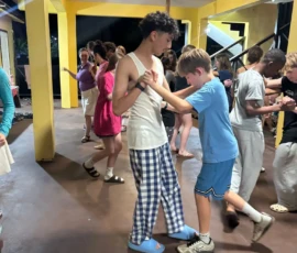 Two boys practice a dance together in a brightly lit room, while other pairs dance in the background. The scene is casual and lively.