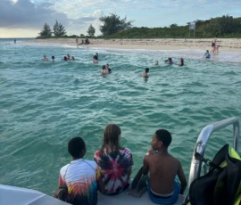 Children sit on a dock looking at people swimming and playing in the shallow turquoise water near a sandy beach with trees in the background.