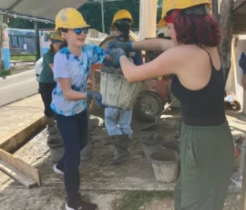 Two people wearing yellow hard hats and gloves pass a bucket between them at a construction site, with others working and buckets visible in the background.
