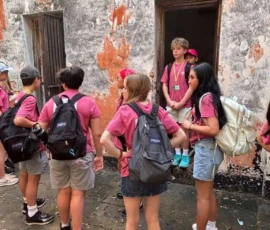 A group of children wearing matching pink shirts and backpacks stands near a weathered wall, some looking toward a doorway where one boy is standing.