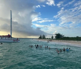 A group of people swims near a catamaran anchored by a small sandy beach, with cloudy skies and the shoreline in the background.