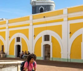 A person with a backpack sits on stone steps in front of a yellow and white colonial building with a lighthouse in the background.