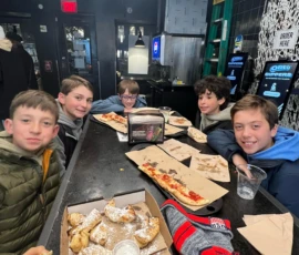 Five boys sit at a table in a casual restaurant with pizza, dessert, and empty plates. The setting appears modern with black walls, a menu screen, and a ladder in the background.