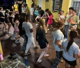 A group of young people in casual clothes gather and dance on a covered patio at night, with some standing and others moving around.