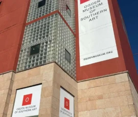 The exterior of the Ogden Museum of Southern Art features large signs with the museum’s name and logo on a red and beige building.