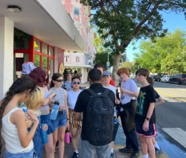 A group of teenagers stands in a circle on a city sidewalk, listening to a man with a black backpack. Some are holding water bottles, and trees line the street.
