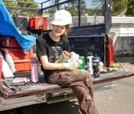 A person wearing a hard hat and work clothes sits on the tailgate of a pickup truck, holding a snack, with tools and water bottles on the truck bed on a sunny day.