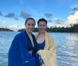 Two young women in swimsuits stand in shallow water, wrapped in blue and yellow towels, with trees and clouds in the background.