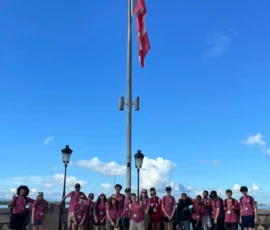 A group of people wearing matching pink shirts stands in front of a large flagpole with a flag, under a blue sky with clouds.