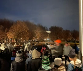 A group of people, including children in winter clothing, view a nighttime outdoor memorial with statues and small American flags. Trees without leaves are visible in the background.