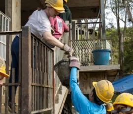 People wearing hard hats work together, passing a bucket up a staircase at a construction or renovation site.