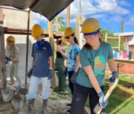 Five people wearing hard hats and gloves work together on a construction site, shoveling and handling buckets of cement under a canopy on a sunny day.