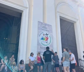 A group of people sits and stands in front of a building entrance with columns and a "Peregrinos de Jesus" sign on the wall. Sunlight creates lens flares in the image.