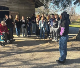 A person stands under a tree speaking to a group of people gathered near a wooden building on a sunny day.