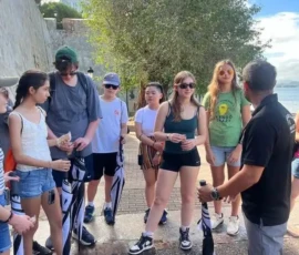 A group of young people with umbrellas stands outdoors near a guide, listening attentively by a tree and a waterfront on a sunny day.
