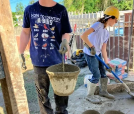 Two people in hard hats work at a construction site; one holds a bucket of wet cement, while the other shovels. Bags of concrete and buckets are visible on the ground.