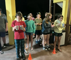 A group of teenagers stands indoors near two orange cones, some holding cups and all dressed in casual clothes, with a nighttime outdoor scene visible behind them.