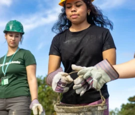 Three people wearing hard hats and gloves work outdoors; the woman in front carries a bucket, with trees and a blue sky in the background.