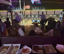A group of people at a bowling alley, some bowling and others seated at tables with food trays and drinks in the foreground.