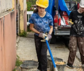 A person wearing a yellow hard hat and blue shirt shovels gravel into buckets on a construction site, with another worker nearby.