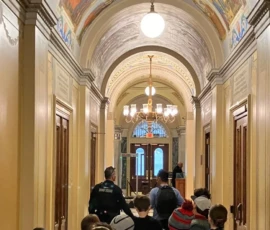 A group of people, including children and two uniformed officers, walk down an ornate hallway with arched ceilings and painted murals.