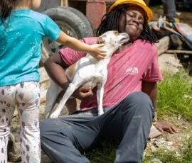 A person in a hard hat sits on the ground, smiling as a dog licks their face. A child standing nearby pets the dog. Outdoor setting with construction materials in the background.