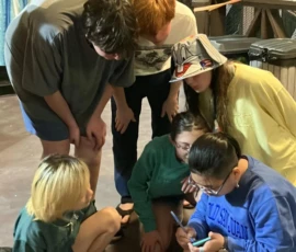 Six people, mostly in casual clothes and sandals, gather closely on a wooden deck at night, looking at a smartphone held by one person sitting on the ground.