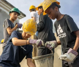 A group of people wearing hard hats and gloves work together outside, passing a bucket as part of a construction or building project.