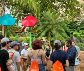 A group of students with orange "The Saklan School" drawstring bags stands in a park near colorful mushroom-shaped structures, listening to an adult.