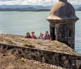 Four people stand and wave near an old stone watchtower overlooking the ocean, with mountains visible in the distance under a cloudy sky.