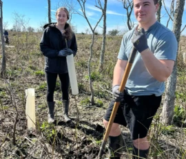 Two people wearing gloves and boots stand in a muddy, leafless area holding tree planting tools and tubes, with young saplings and trees visible around them under a blue sky.