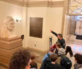 A group of students listens to a guide near a large bust of Abraham Lincoln and a plaque in a building hallway.