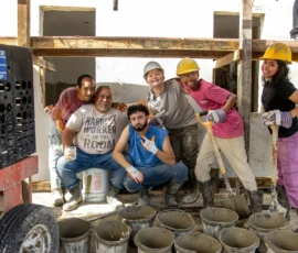 A group of six people wearing work clothes and helmets pose and smile at a construction site with buckets of cement in front of them.