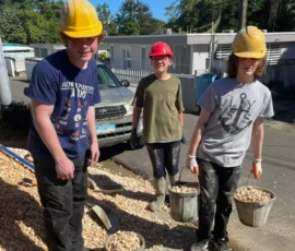 Three young people wearing hard hats carry buckets filled with rocks at a construction site on a sunny day.