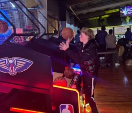 A boy plays an arcade basketball game indoors, aiming to shoot a basketball; several people are seated in the background.