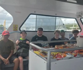 Six boys sit on a boat next to a snack counter, with docks and water visible through the window under a cloudy sky.
