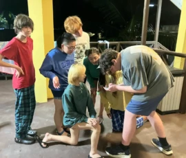 A group of seven kids gather closely together on a covered patio at night, looking intently at something on a phone held by one of them.