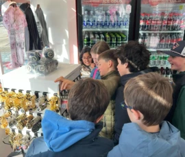 A group of children in jackets look at a spinning rack of souvenirs inside a store with refrigerated drinks in the background.
